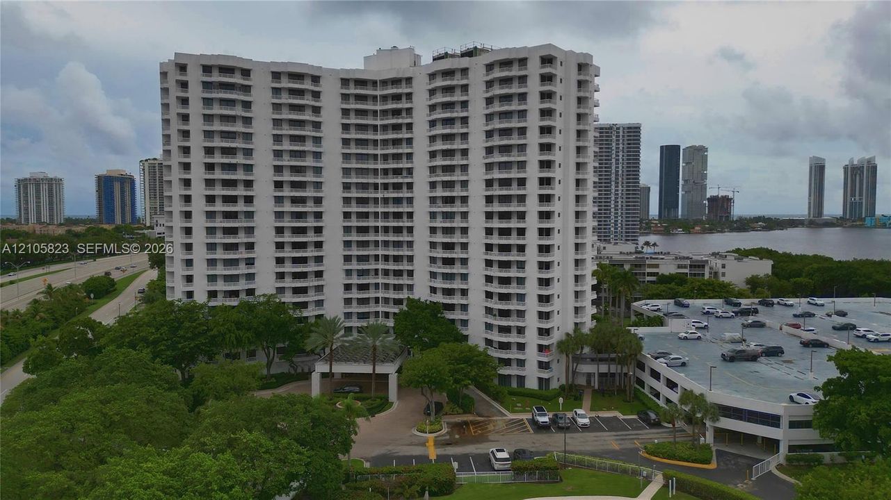 High-rise residential building featuring a curved architectural design, multiple private balconies, a covered arrival portico, and an integrated multi-level parking structure
