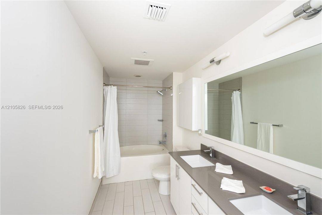 Bathroom featuring a dual-sink vanity with a solid-surface countertop, full-width wall mirror, horizontal wall-mounted vanity lighting, and a tiled tub-shower enclosure