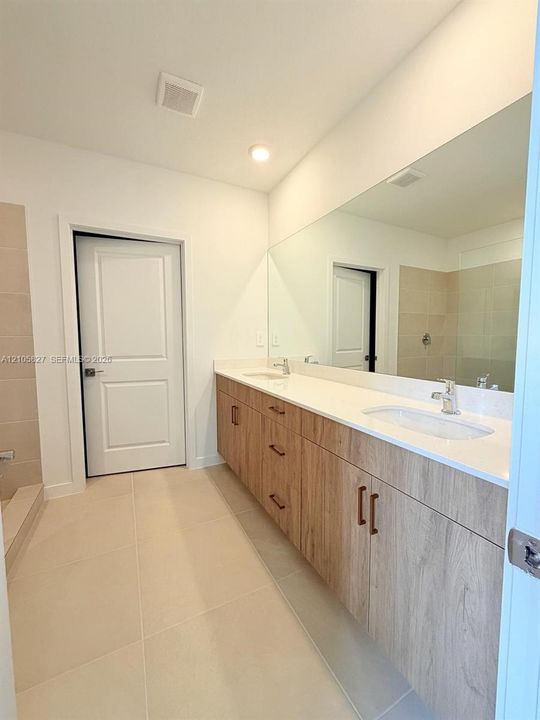 Double vanity featuring wood-finish cabinetry, solid-surface countertops, recessed sinks, and oversized wall-to-wall mirror with neutral-toned floor and shower tile