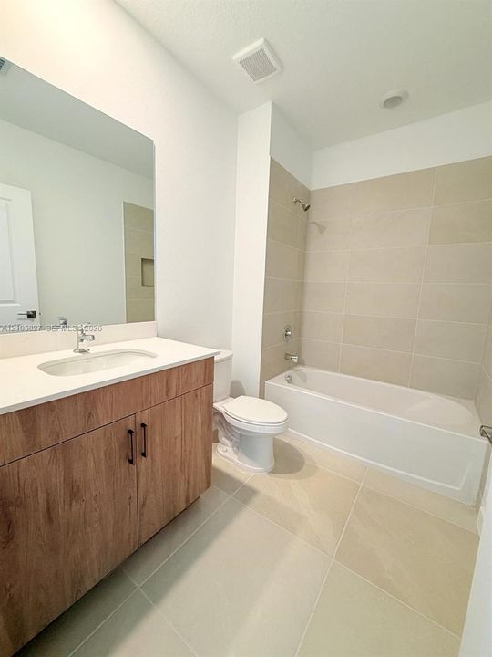 Bathroom featuring a wood-finish vanity with a light-toned countertop, large-format neutral floor tiling, a porcelain-enamel bathtub, and full-height tile shower surround