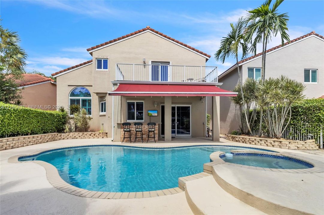 Stucco exterior featuring an in-ground swimming pool, integrated spa, retractable patio awning, and a second-story balcony with metal railing