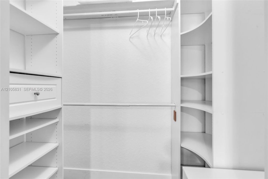 Walk-in closet featuring white custom built-in shelving, curved corner display units, dual-level hanging rods, and a soft-close drawer