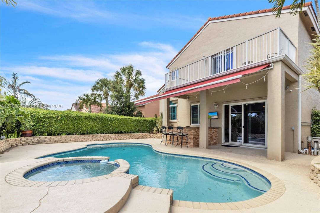 Backyard area featuring an in-ground swimming pool, attached spa, stone-veneer outdoor bar, retractable awning, and second-story balcony with metal railing
