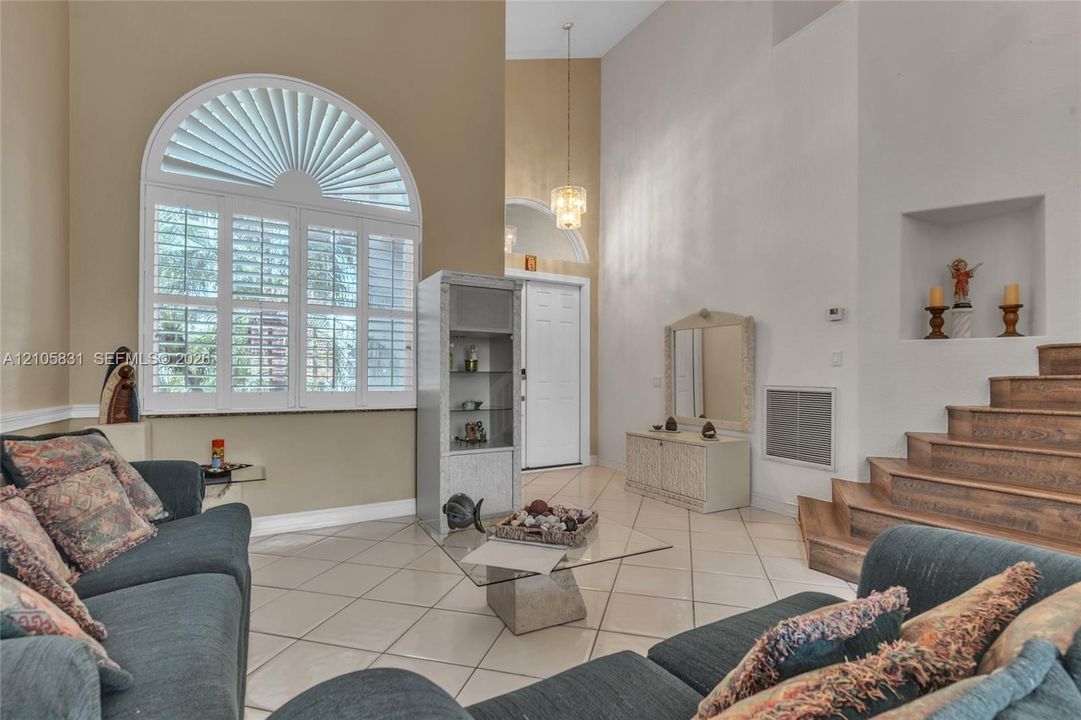 High-ceilinged living space featuring an arched window with plantation shutters, tile flooring, a recessed wall niche, and wood-finish stairs