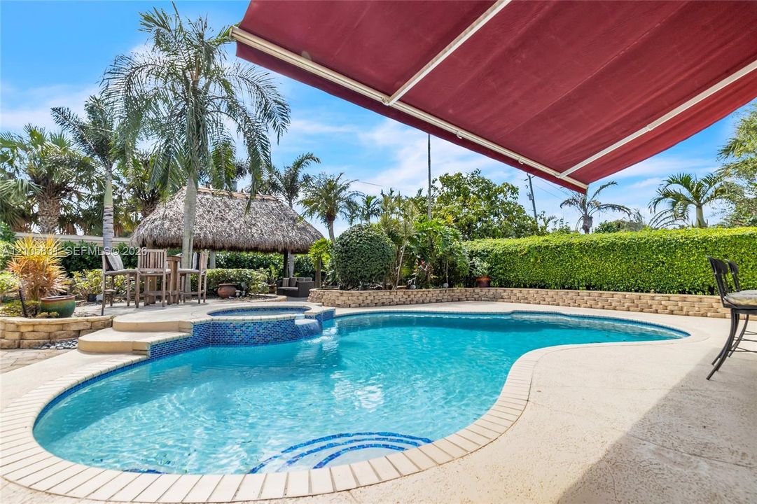 In-ground pool with stone coping and integrated spa, retractable awning, detached thatch-roofed cabana, and tiered stone retaining walls