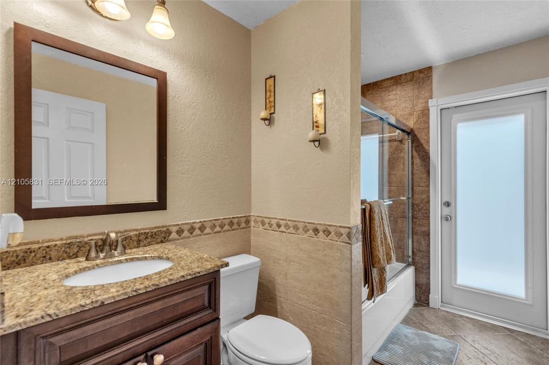 Bathroom featuring a wood-finish vanity with a stone-composite countertop, integrated sink, framed wall mirror, half-wall tile wainscoting with decorative accent border, and a glass-enclosed shower