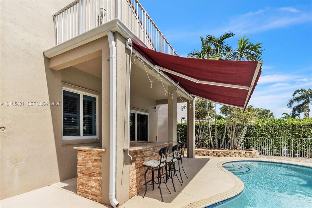 Covered outdoor patio featuring a stacked stone bar, retractable fabric awning, second-level balcony with white railings, and swimming pool