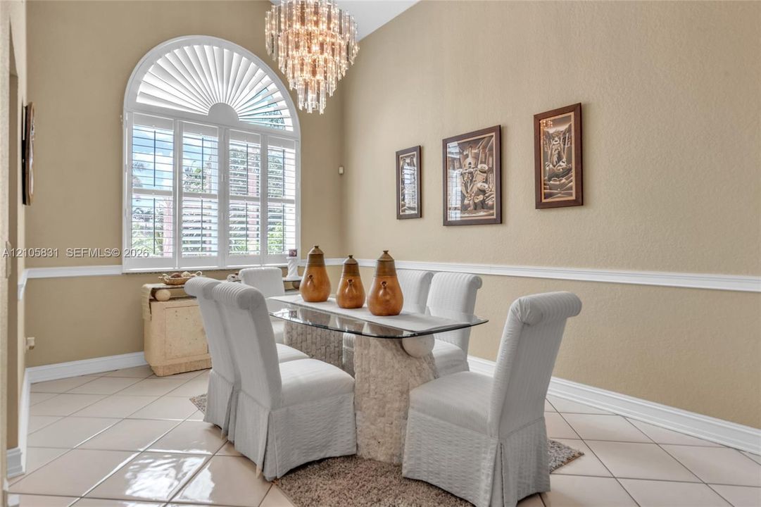 High-ceilinged room featuring an arched window with plantation shutters, tile flooring, crown molding, and a tiered crystal chandelier