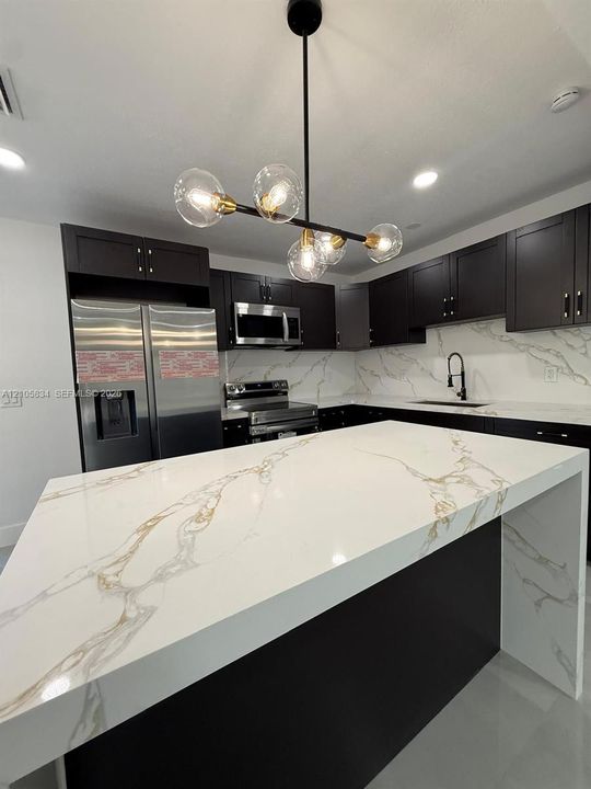 Kitchen featuring an expansive waterfall island with a veined stone-finish countertop, dark cabinetry, stainless steel appliances, and a multi-bulb statement chandelier
