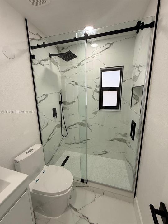 Walk-in shower featuring large-format wall tile, glass sliding door with matte black hardware, rainfall showerhead, integrated recessed shelf, and matte black plumbing fixtures