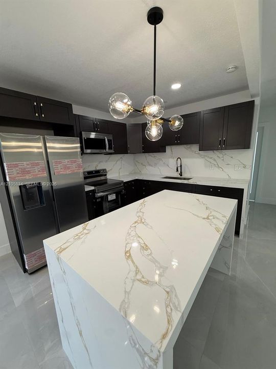 Kitchen featuring dark cabinetry, expansive waterfall-edge island, stone-patterned countertops with matching backsplash, and a contemporary multi-globe pendant light fixture