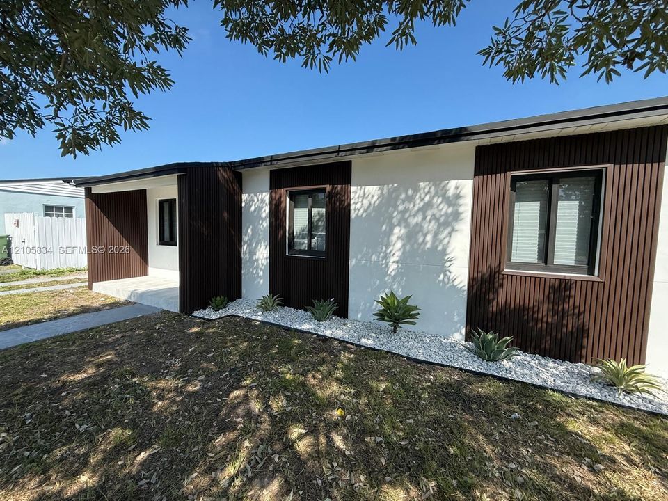 Exterior facade featuring white stucco walls and dark-toned vertical corrugated metal siding, complemented by dark-framed windows and a stone-covered landscape bed