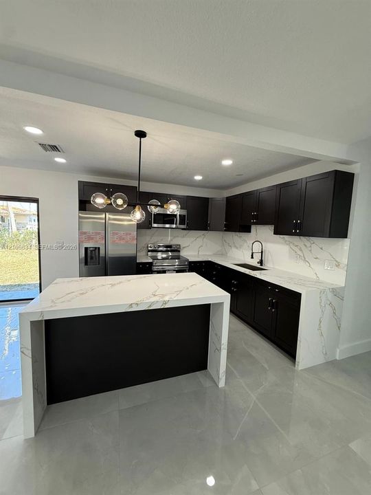 Kitchen featuring dark cabinetry, stone-finish waterfall island, stone-finish backsplash, matte black hardware, and pendant lighting