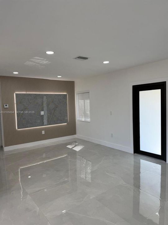 Polished large-format tile flooring, accent wall featuring vertical slat paneling with integrated lighting, centered stone-finish panel, and a black-framed glass door