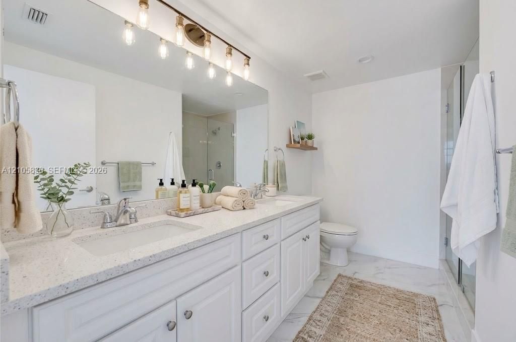 Dual-sink vanity with white cabinetry, light-toned stone-finish countertops, polished chrome fixtures, and a large-scale mirror with an integrated multi-bulb light fixture