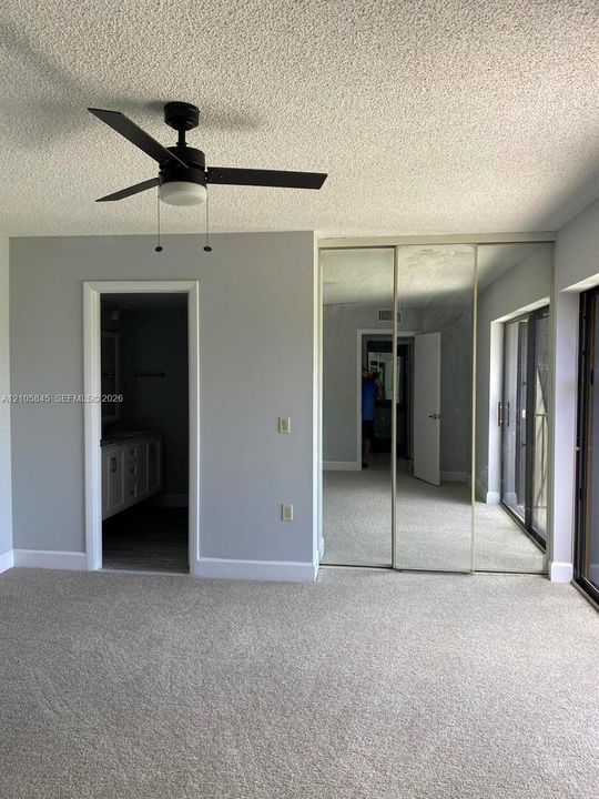 Neutral-toned interior featuring wall-to-wall carpeting, three-panel mirrored closet doors, a ceiling fan with integrated lighting, and a sliding glass door