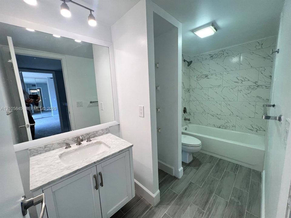 Bathroom featuring a vanity with stone-finish countertop, undermount sink, full-width mirror, tile-surround bathtub, and wood-finish flooring