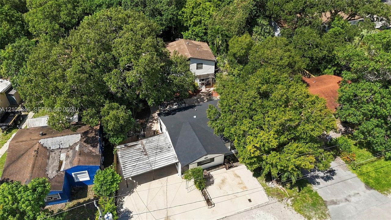 Aerial view featuring a multi-level structure with a dark shingled roof, a concrete driveway, and an attached covered carport, situated among mature trees
