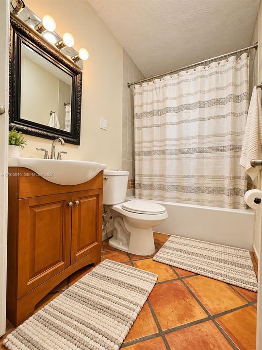 Bathroom featuring a warm-toned wood-finish vanity with a curved basin, rectangular wall-mounted mirror with a decorative frame, three-bulb vanity light fixture, and terra-cotta tile flooring