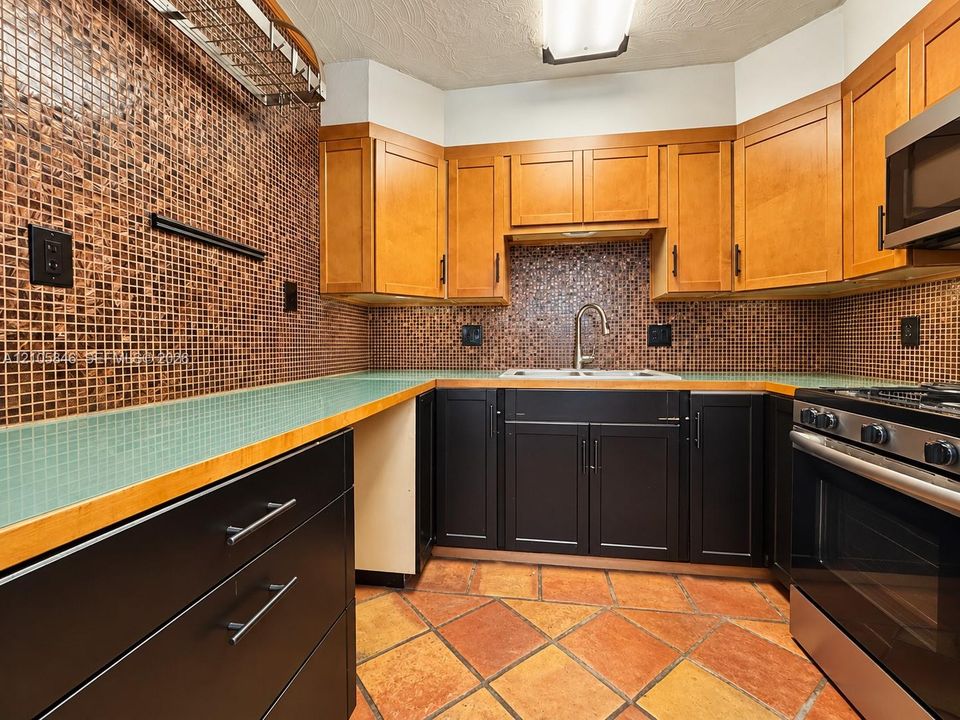 Kitchen featuring two-tone cabinetry, mosaic tile backsplash, solid surface countertops, a stainless steel gas range, and terracotta tile flooring