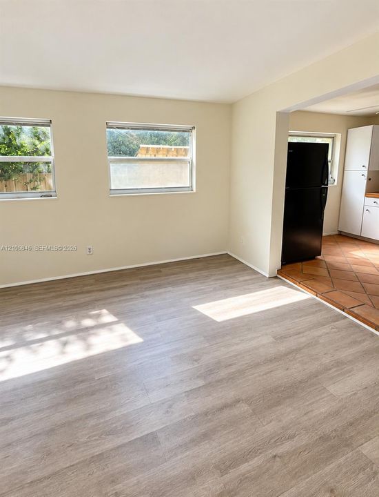 Neutral interior space featuring wood-finish flooring, dual windows, and terra-cotta tile flooring in the kitchen area with white cabinetry and a black refrigerator