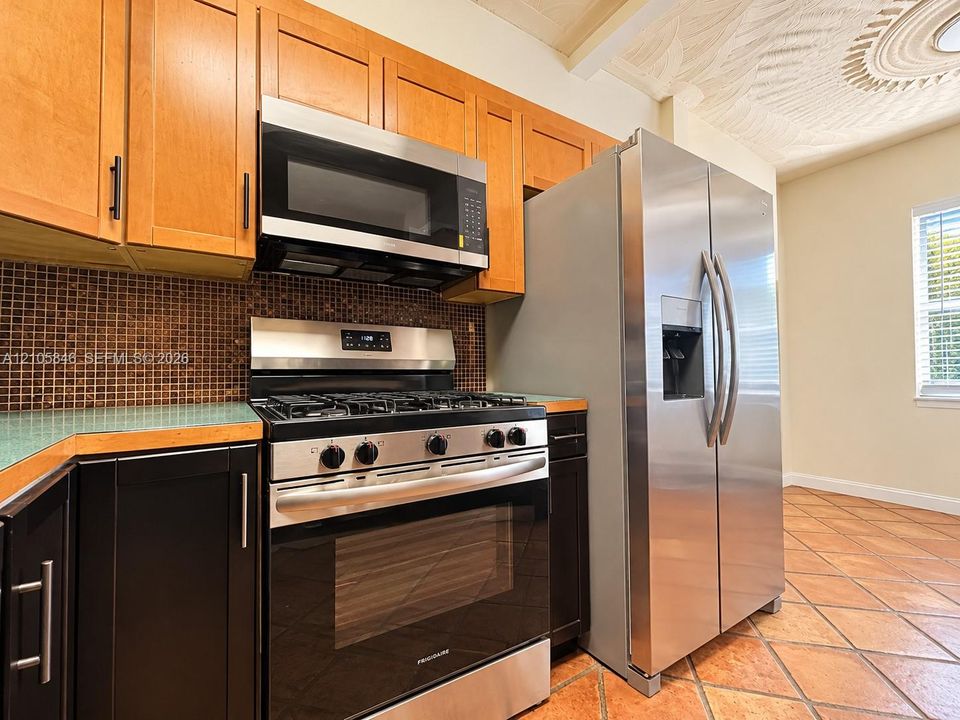 Kitchen featuring two-tone cabinetry, stainless steel gas range and refrigerator, mosaic tile backsplash, and terracotta tile flooring
