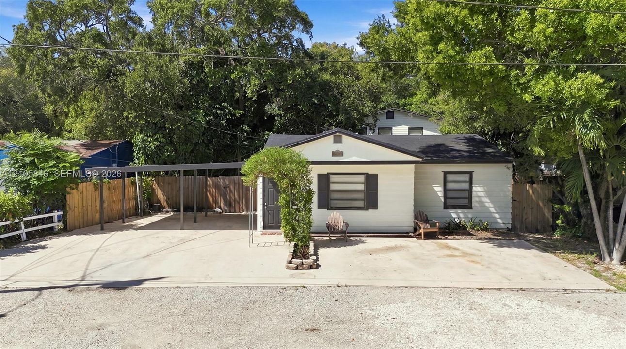 Single-story residence featuring light-colored horizontal siding, dark-toned shutters, an attached metal-roofed carport, a spacious concrete driveway, and surrounding mature trees