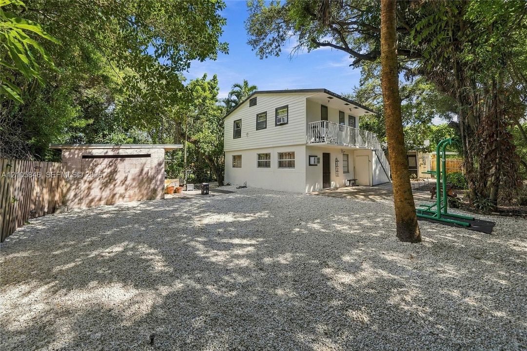 Two-story exterior with horizontal siding, gravel ground cover, detached concrete block structure, second-level balcony with metal railing, and mature trees