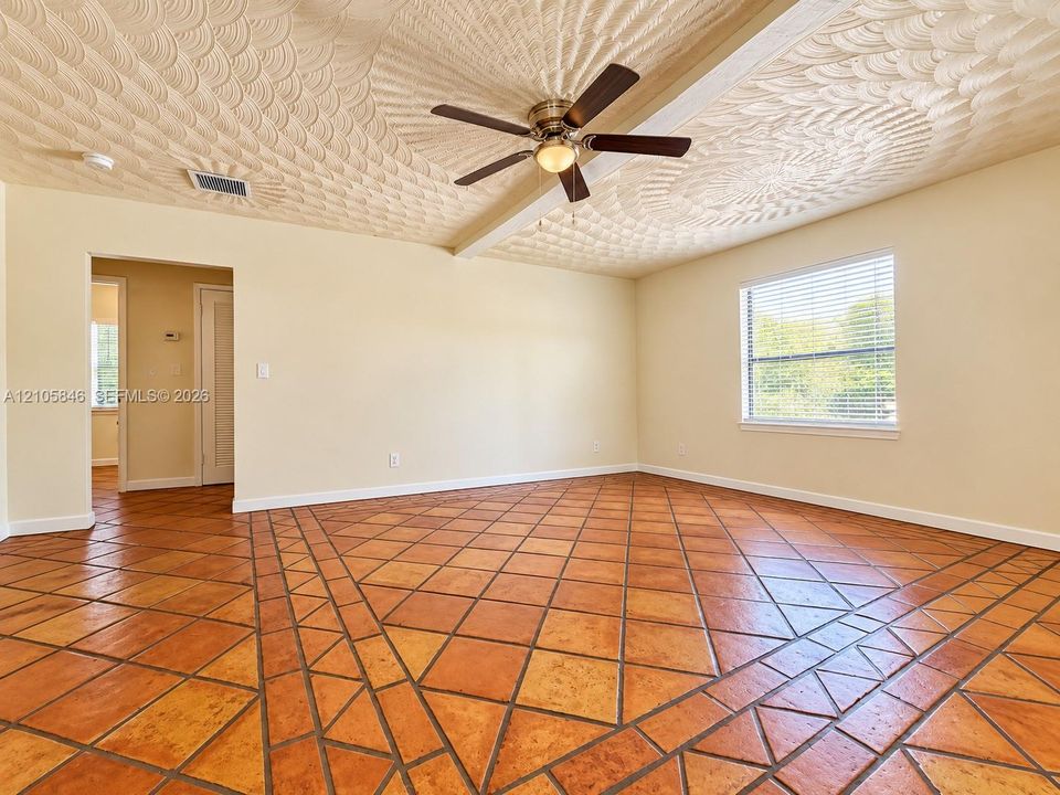 Textured ceiling featuring a central ceiling fan with integrated lighting, clay tile flooring in a geometric pattern, and a window with horizontal blinds