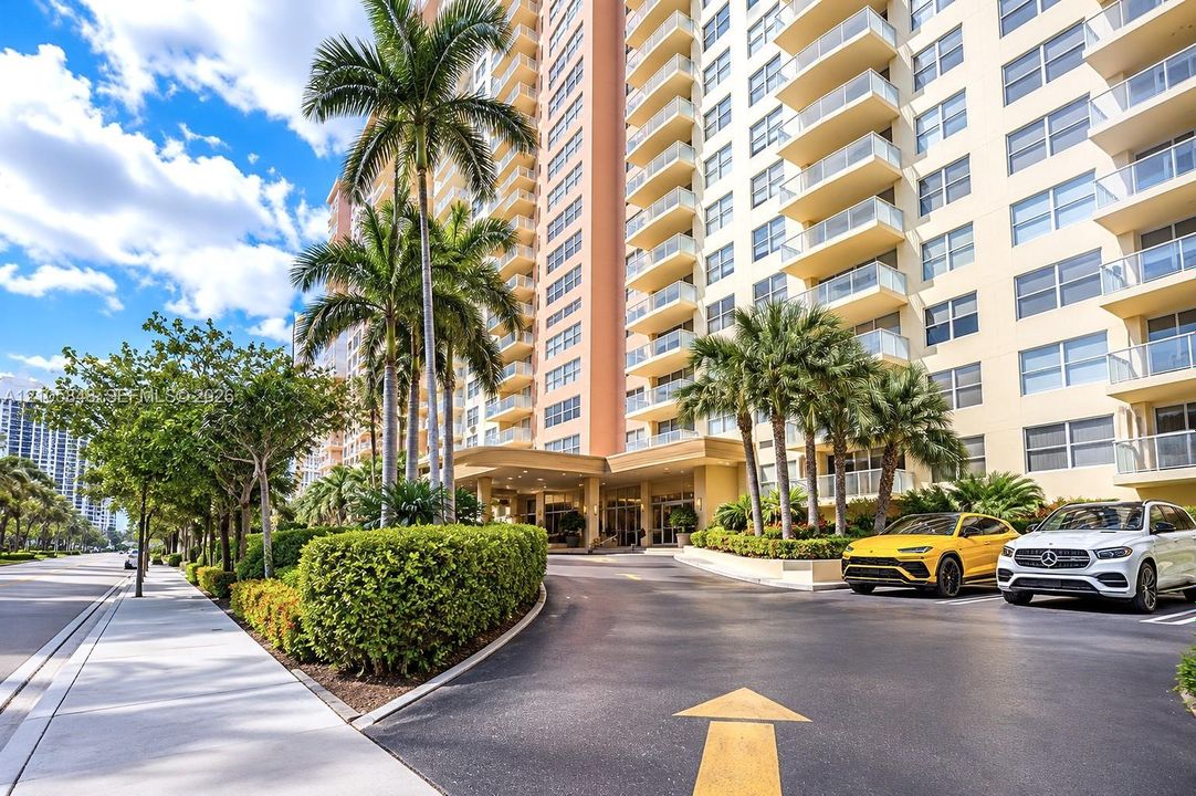 High-rise building exterior featuring a covered porte-cochere, glass-railed private balconies, mature palm trees, and manicured shrubbery