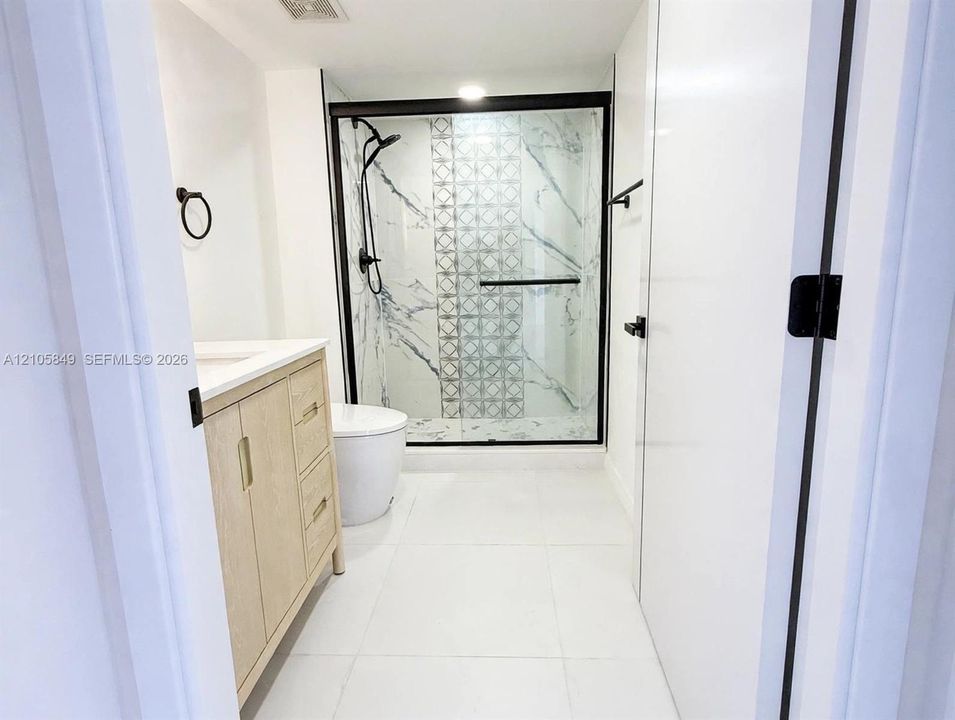 Bathroom featuring a light wood-finish vanity, large-format tile flooring, and a glass-enclosed shower with decorative accent tile, stone-patterned wall panels, and matte black fixtures