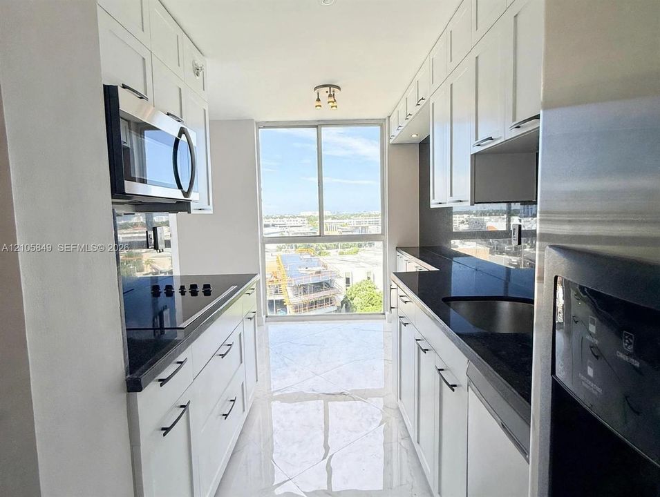 Galley kitchen featuring white Shaker-style cabinetry, dark stone countertops, stainless steel appliances, large-format tile flooring, and floor-to-ceiling windows