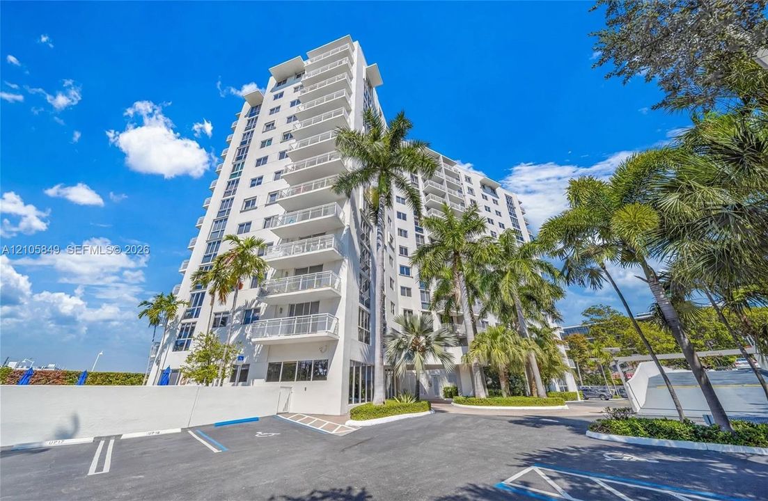 High-rise residential building featuring white stucco facade, stacked balconies, and paved surface parking, set amidst mature palm trees and manicured landscaping