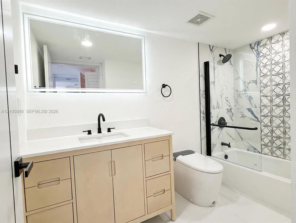 Bathroom featuring a light wood vanity with a white countertop, wall-mounted LED-backlit mirror, matte black fixtures, and a glass-enclosed tub with patterned tile detailing