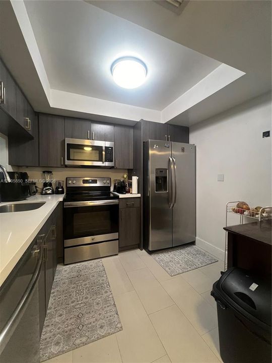 Kitchen featuring dark-toned cabinetry, stainless steel appliances, solid-surface countertops, tile flooring, and a tray ceiling