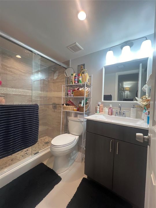Walk-in shower with glass enclosure, decorative stone-finish tile surround, dark wood-tone vanity cabinet, rectangular under-mount sink, and three-light wall fixture