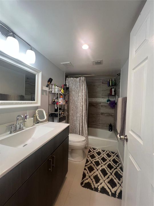 Dark finish vanity with a rectangular integrated sink, quartz countertop, polished chrome faucet, multi-light vanity fixture, and tiled tub-shower surround with a decorative mosaic accent strip