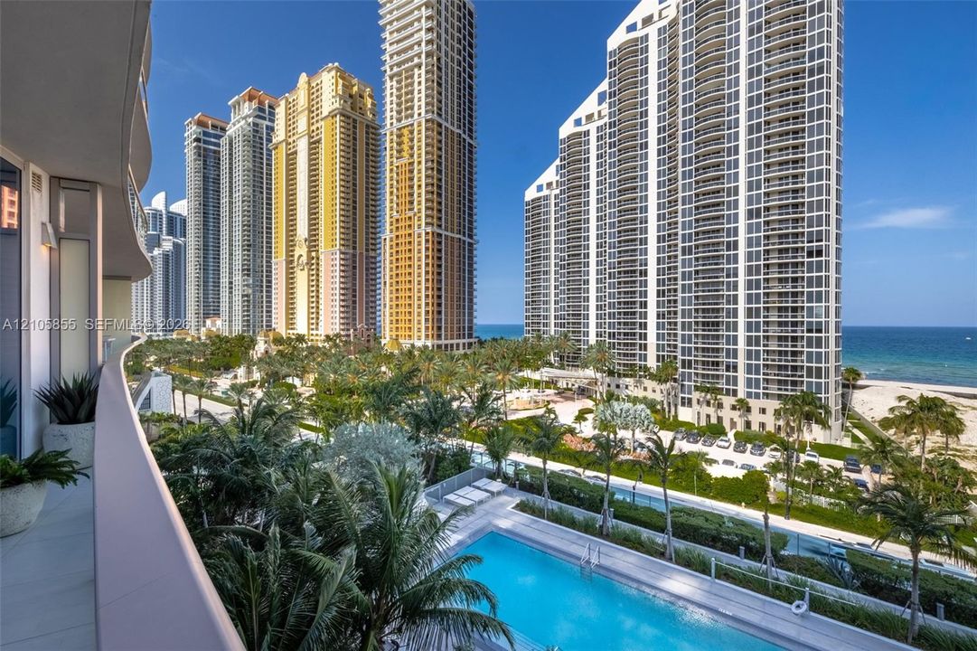 Curved balcony with tile flooring, overlooking an outdoor swimming pool, palm-lined grounds, high-rise buildings, and the ocean