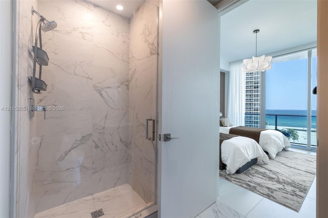 Walk-in shower featuring large-format stone-tile surround, glass enclosure, and ceiling-mounted light, accompanied by floor-to-ceiling windows and coastal water scenery