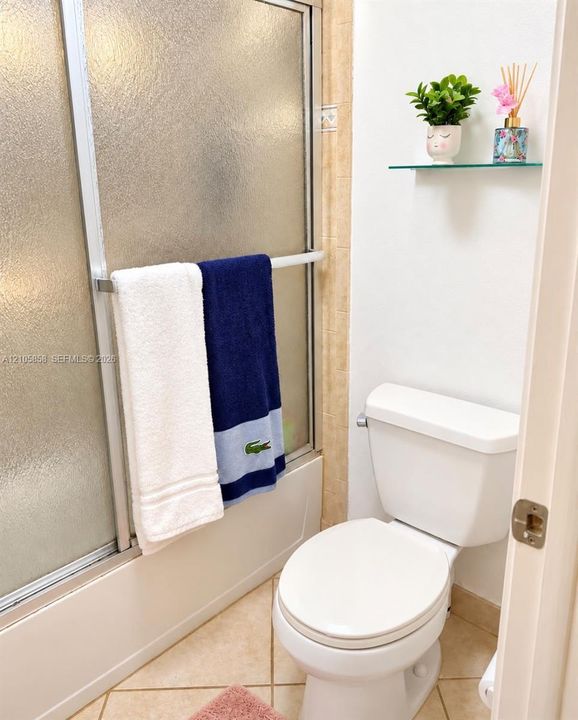 Shower enclosure with textured glass doors, white porcelain toilet, and wall-mounted glass shelving unit featuring tile flooring