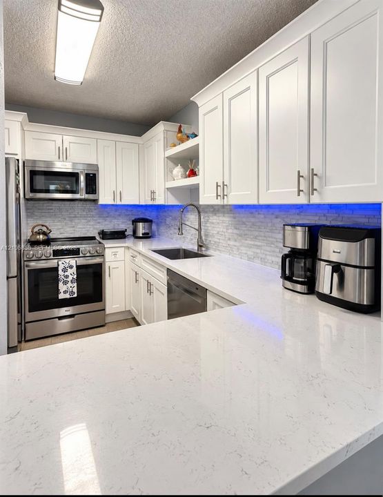 Kitchen featuring white shaker-style cabinetry, solid-surface countertops, a stainless steel appliance package, and a tiled backsplash with integrated under-cabinet lighting