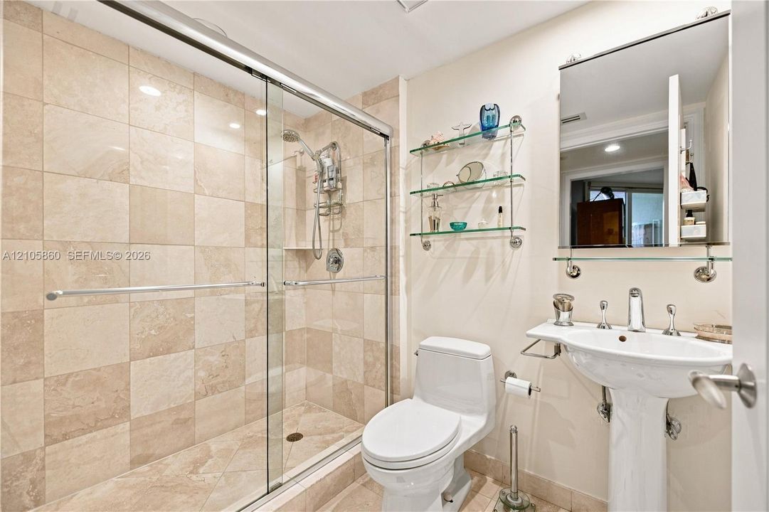 Bathroom featuring a glass-enclosed walk-in shower with stone-finish tile surround, pedestal sink with polished metal fixtures, and built-in glass shelving