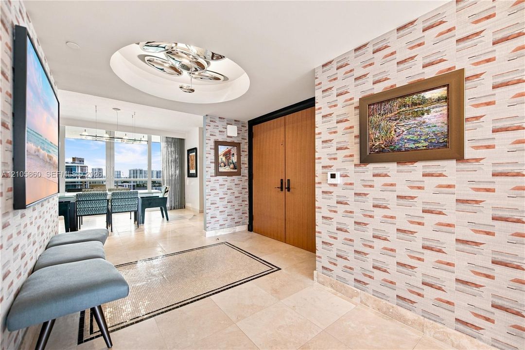 Foyer featuring patterned wallpaper, stone tile flooring with a decorative mosaic inlay, recessed circular ceiling detail with multi-bulb lighting, and wood-finish double doors