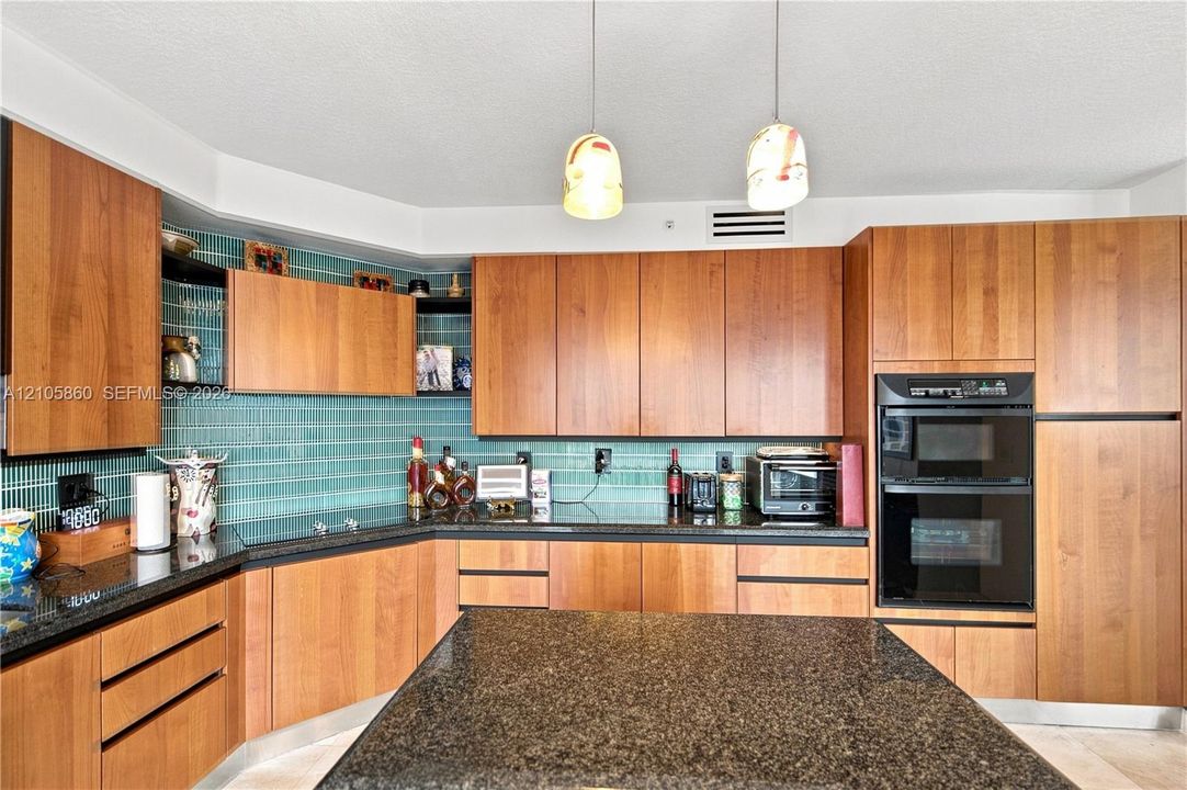 Kitchen featuring flat-panel wood cabinetry, dark stone countertops, a green glass tile backsplash, double wall ovens, and dual pendant lighting