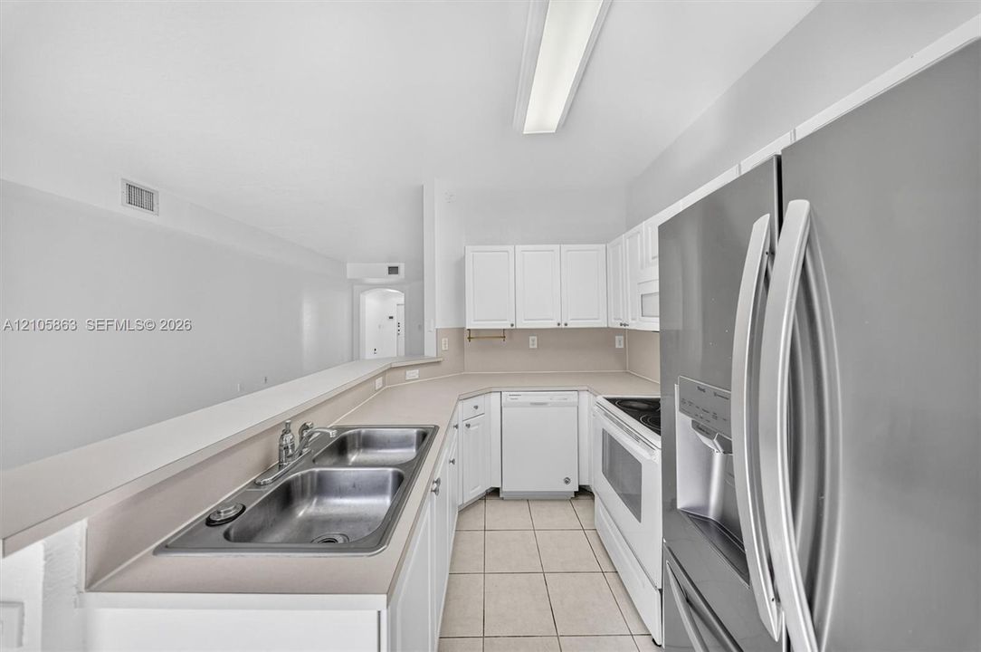 Kitchen featuring white cabinetry, solid-surface countertops, dual-basin stainless steel sink, tile flooring, and stainless steel refrigerator