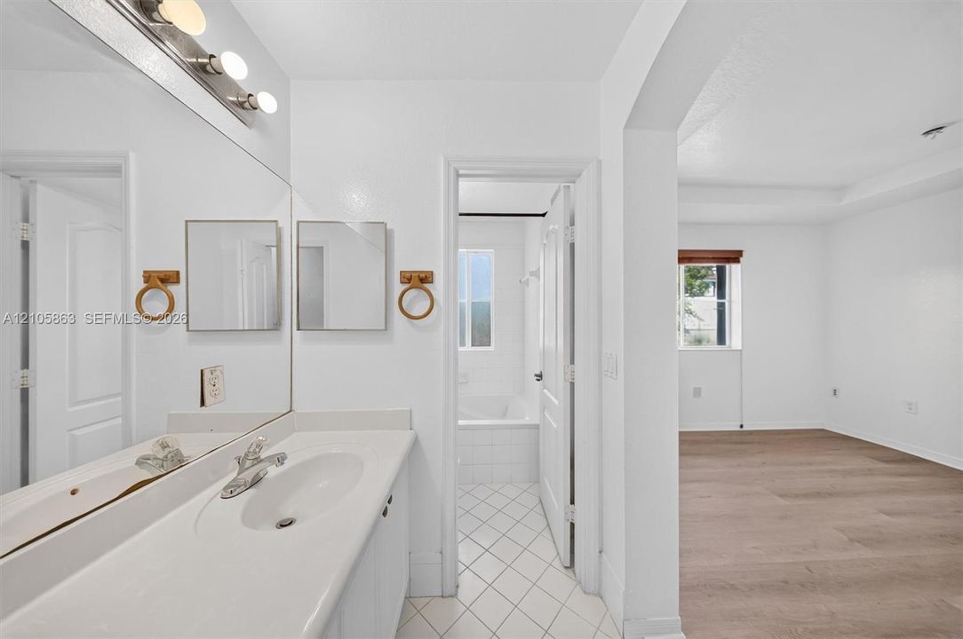 Full-width vanity with integrated sink, expansive wall-to-wall mirror, brushed metal fixtures, white tiled flooring, and wood-finish flooring