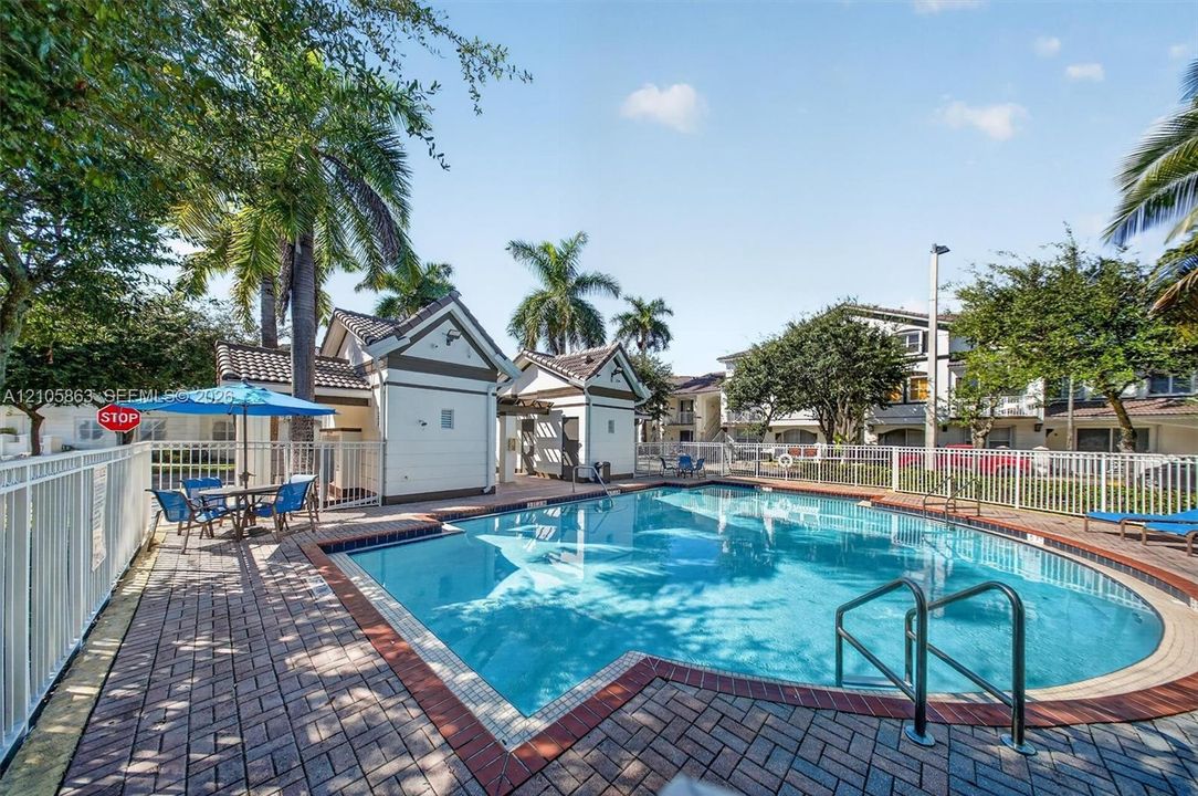 In-ground swimming pool with brick paver decking, white perimeter fencing, and two pool-side structures featuring pitched rooflines