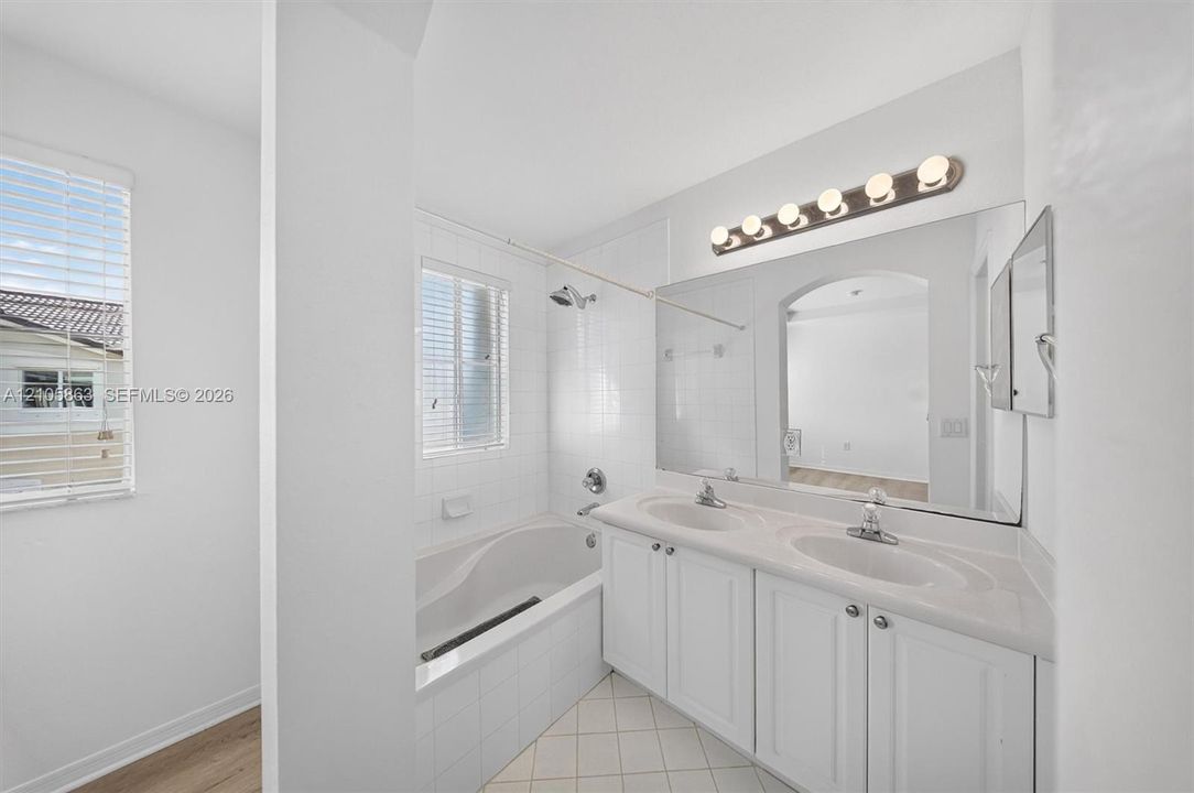 Dual-sink vanity with white cabinetry and a solid-surface countertop, full-width wall mirror, bathtub with white tile surround, and tile flooring