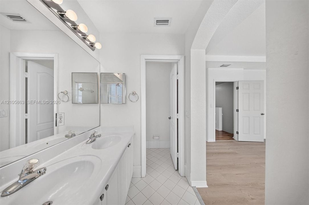 Dual-sink vanity with white countertop, full-width vanity mirror, polished chrome fixtures, white tile flooring, and arched architectural detailing