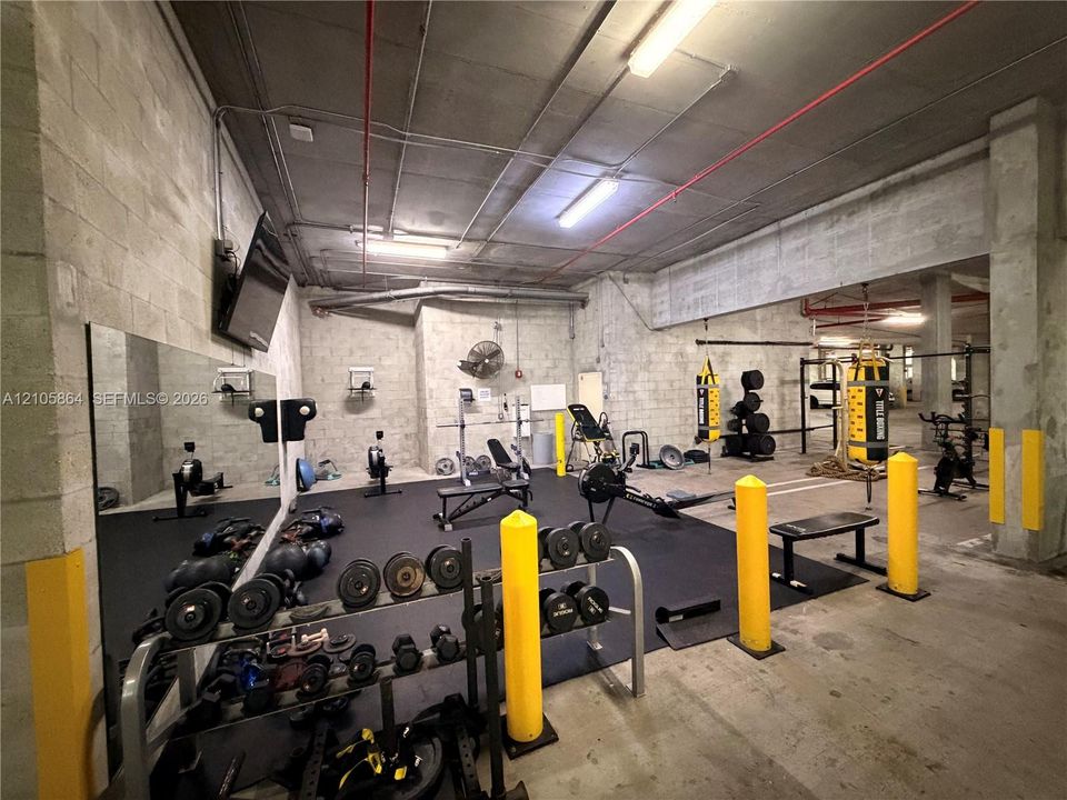 Fitness area featuring exposed concrete ceiling, industrial-style lighting, large wall-mounted mirror, and impact-resistant rubber flooring